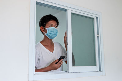 Close-up of mature man wearing flu mask holding phone seen through window