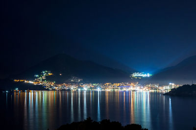 Illuminated city by lake against sky at night