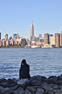 Rear view of man sitting on rocks by river against cityscape