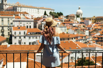 Rear view of young woman looking at buildings while standing by railing in balcony against clear sky