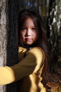 A girl in a yellow sweater hugs a tree in the forest