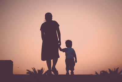 Silhouette of mother and son holding hands while standing against orange sky