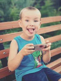 Portrait of boy with sticking out tongue holding mobile phone while sitting on bench
