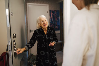 Smiling senior woman opening door while female nurse arriving at home