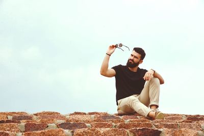 Full length of man sitting on steps while holding sunglasses against sky