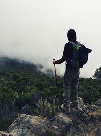 Rear view of hiker standing on rock at mountain against sky during foggy weather