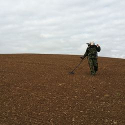 Low angle view of army soldier using metal detector on field against cloudy sky