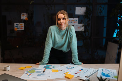 Smart blonde designer looking at the camera and posing leaning at the table at the evening office