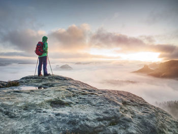 Tourist in green windcheater with hood on head and red backpack on back enjoy wild misty viee