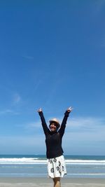 Teenage girl with arms raised standing by sea against blue sky during sunny day