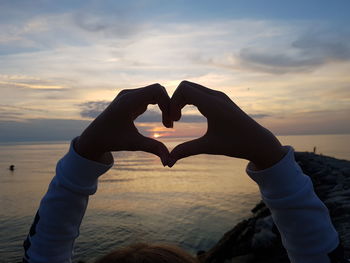 Hand holding heart shape against sea during sunset