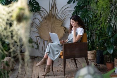 Thoughtful woman sitting in chair in home garden, working on laptop surrounded by exotic houseplants