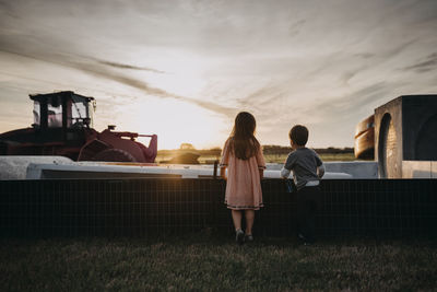 Rear view of siblings standing on grassy field at farm against sky during sunset