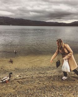 Smiling woman feeding ducks at lake against sky