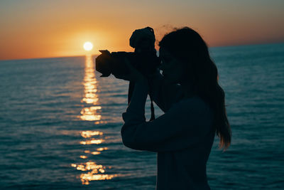 Silhouette woman photographing sea against sky during sunset