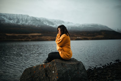 Man sitting on rock by lake against mountains