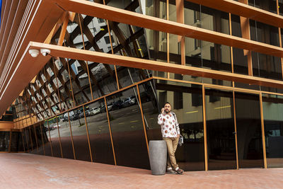 Casual wear man standing against office building while using mobile