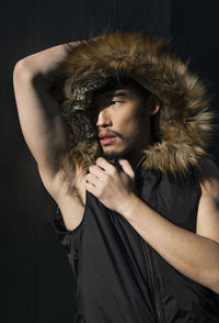 Young man wearing hooded jacket looking away while standing against black background