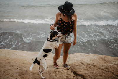 Unrecognizable trendy young lady in black hat and casual clothes along rocky coast with obedient dappled dog against troubled sea water