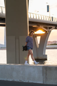 Freedom lifestyle. woman of middle age walk near river hold longboard over big city view at sunset