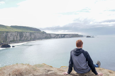 Rear view of teenage boy sitting on cliff against sea
