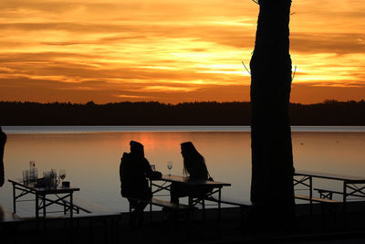 Silhouette people sitting by lake against sky during sunset