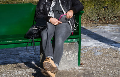 Low section of woman using mobile phone while sitting on bench during winter