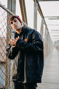 Portrait of young man standing by chainlink fence outdoors