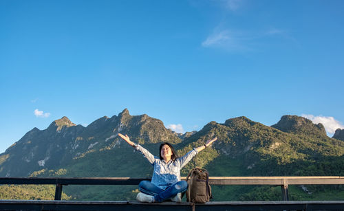 Woman with arms outstretched sitting by railing against blue sky