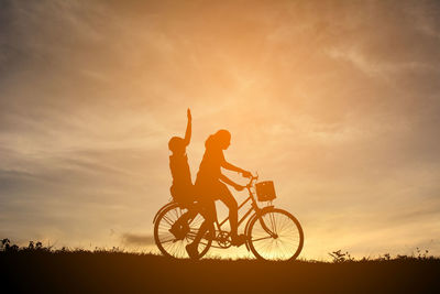 Silhouette siblings riding bicycle on field against sky during sunset