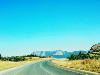 Empty road by mountains against clear blue sky