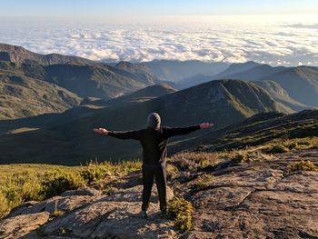 Rear view of man with arms outstretched standing on mountain against sky