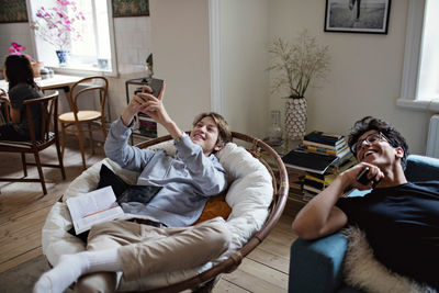 Smiling teenage boy taking selfie while studying with friend in living room at home