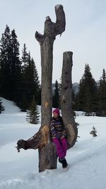 Smiling girl sitting on tree trunk at snow covered field against sky