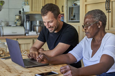 Senior woman and smiling man sitting at kitchen table sharing laptop