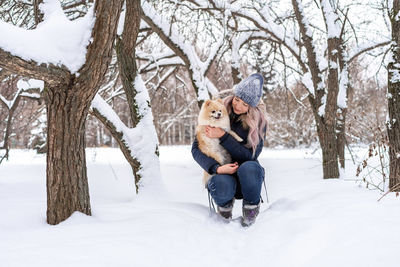 Young woman carrying dog while crouching in snow during winter