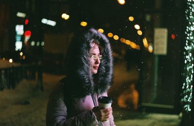 Young woman wearing warm clothing holding disposable cup while standing outdoors at night