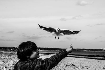 Side view of boy feeding bird at beach against sky