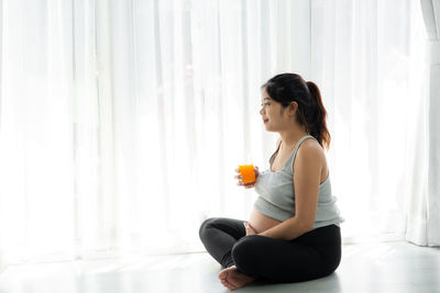 Side view of pregnant woman holding juice while sitting on floor at home