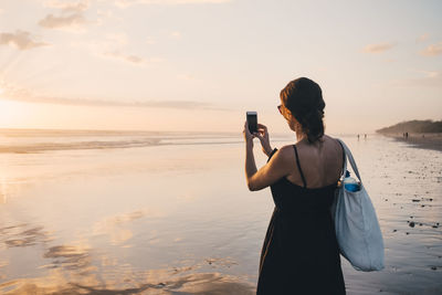 Rear view of woman photographing sunset over sea using mobile phone at beach