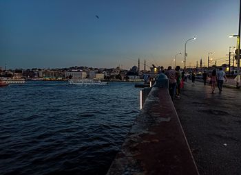 People on sea by buildings against sky at sunset