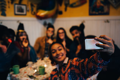 Smiling young woman taking selfie with multi-ethnic friends holding props while sitting at restaurant during dinner part