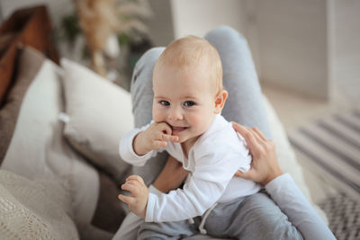 The baby, 10 months old, gnaws a finger sitting on mum's hands, white bodysuit. cozy real interior 