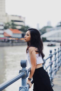 Side view of young woman standing at railing by river in city