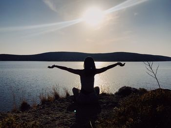 Rear view of silhouette woman with arms outstretched sitting by lake