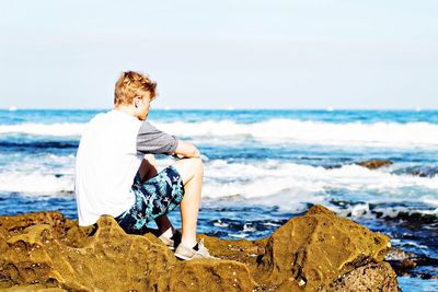 Man sitting on rock by sea against sky during sunny day