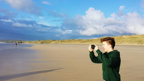 Young man photographing with smart phone at beach against sky