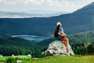 Full length of young woman sitting on rock against mountains