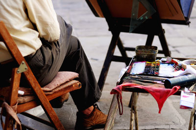 Low section of man sitting with art and craft equipment on table at footpath