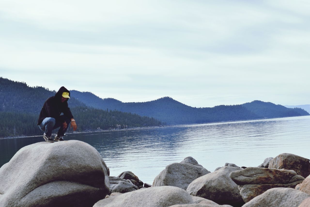 water, mountain, sky, men, lifestyles, leisure activity, rear view, rock - object, standing, scenics, mountain range, tranquil scene, lake, tranquility, nature, looking at view, sitting, beauty in nature